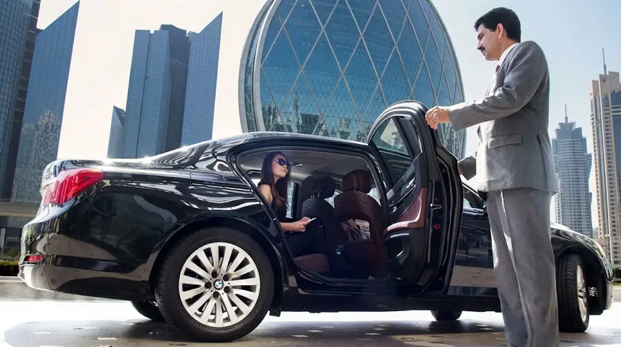 Professional chauffeur opening the door of a black luxury BMW for a passenger in downtown Abu Dhabi with iconic skyline.