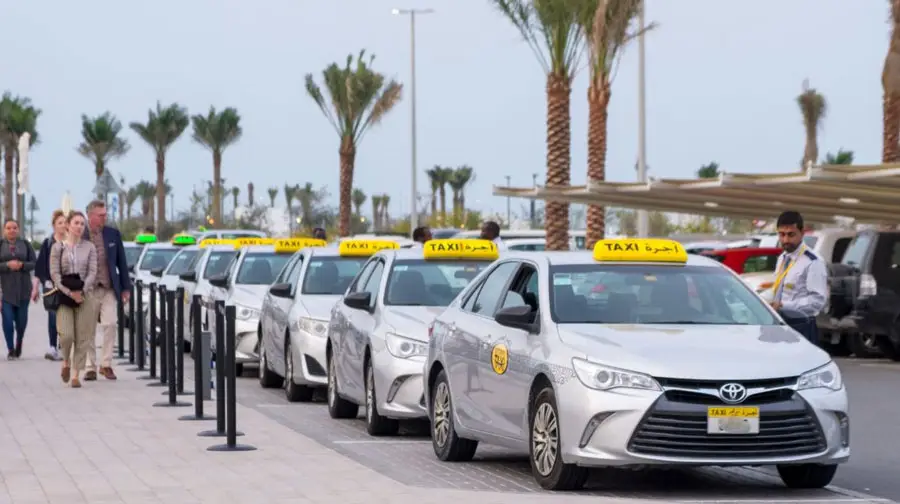Row of silver Abu Dhabi taxis with yellow roof signs lined up at a terminal with palm trees and passengers nearby.