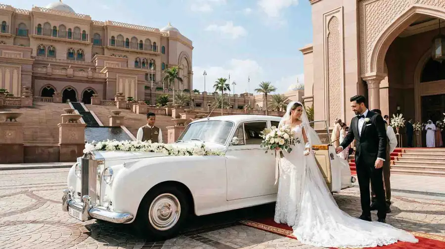 A bride and groom arriving in a classic white vintage Rolls Royce decorated with flowers at a grand palace, illustrating a cost guide for wedding transportation services in Abu Dhabi.