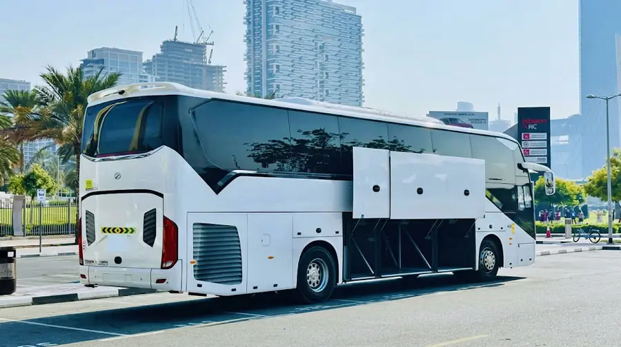 White luxury coach with open luggage compartments parked near modern high-rises and palm trees in Abu Dhabi.