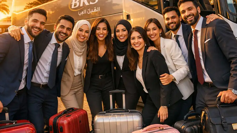 A diverse group of smiling business professionals in formal attire and hijabs posing together with their luggage in front of a luxury shuttle bus at Abu Dhabi airport.