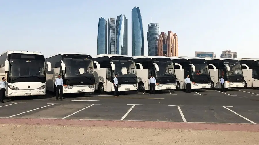 A fleet of white luxury charter buses parked in a lot with drivers standing alongside, set against the Abu Dhabi skyline featuring Etihad Towers.