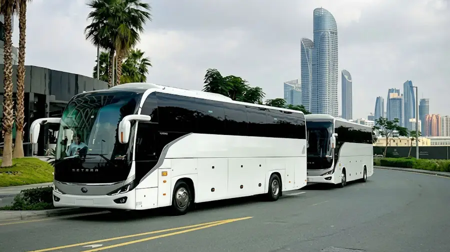 Two modern white passenger coaches driving along a road lined with palm trees, with high-rise skyscrapers in the background of Abu Dhabi.
