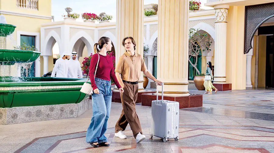 Young couple with rolling luggage walking past ornate columns and a fountain outside a luxury Abu Dhabi hotel.